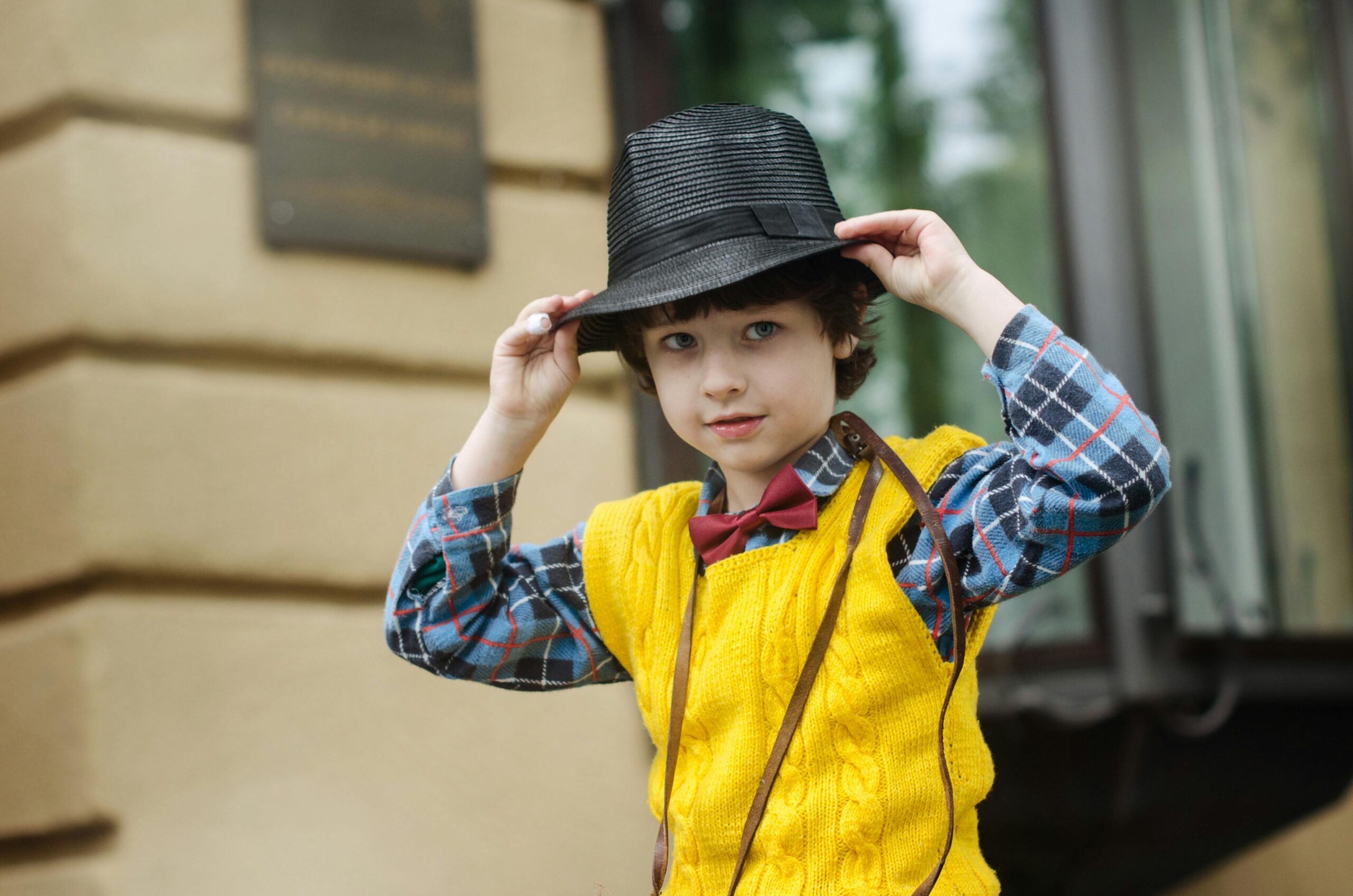 A stylish young boy wearing a yellow sweater, plaid shirt, and black hat poses playfully outdoors.
