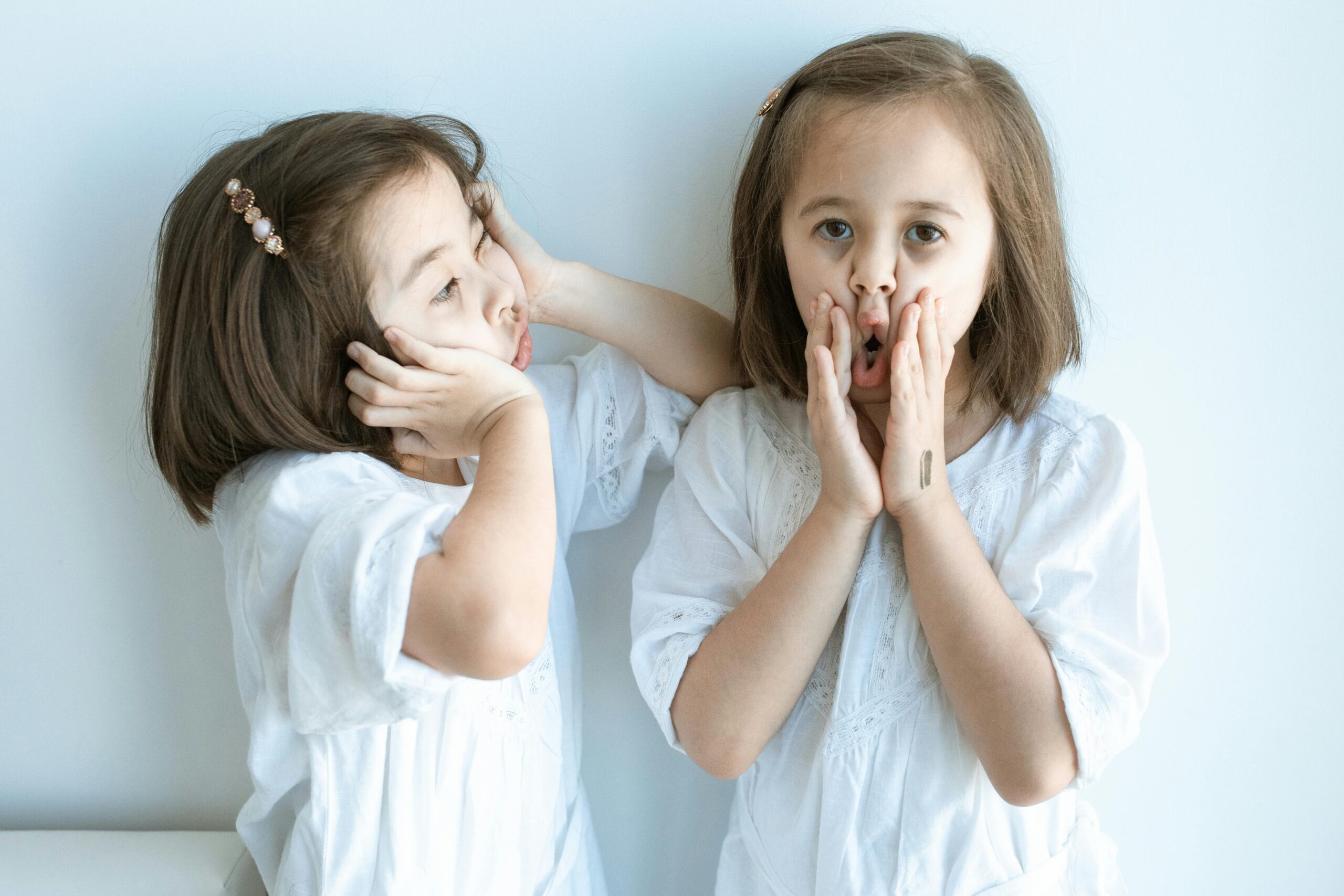 Two young girls in white dresses express playful emotions indoors against a light background.